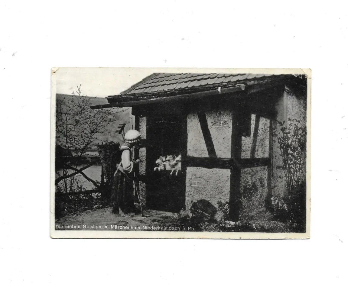 „schwarz-weiß bedruckte Ansichtskarte aus den 1960er Jahren mit Märchenszene aus dem Märchenhain Niederheimbach am Rhein. Motiv zeigt Figuren aus der Wolf und die sieben die sieben Geisslein - Abschied der Mutter und zeigt ein Haeusschen mit dekorativen Elementen.“