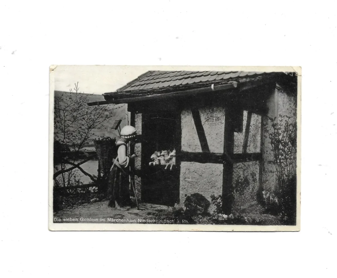 „schwarz-weiß bedruckte Ansichtskarte aus den 1960er Jahren mit Märchenszene aus dem Märchenhain Niederheimbach am Rhein. Motiv zeigt Figuren aus der Wolf und die sieben die sieben Geisslein - Abschied der Mutter und zeigt ein Haeusschen mit dekorativen Elementen.“