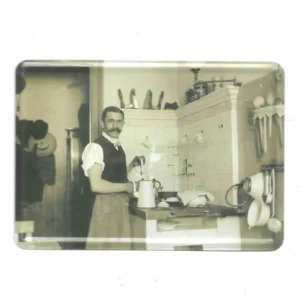 „Blechpostkarte mit Schwarz-Weiß-Foto: Mann in historischer Küche mit Töpfen, Schürze und Kochgeschirr, ca. 10 × 14 cm.“
