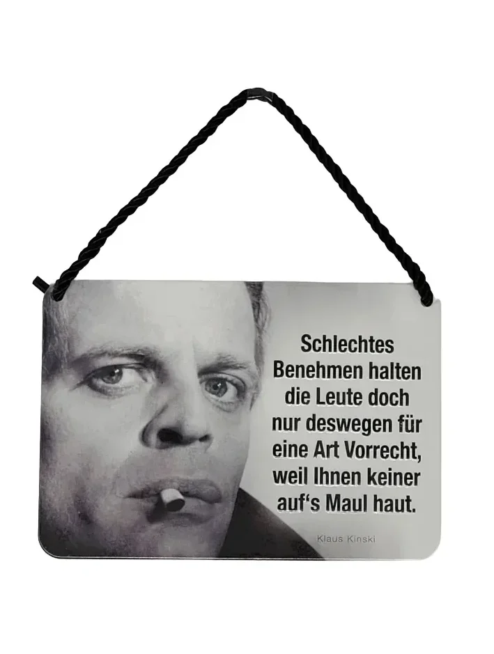 „Blechpostkarte mit Schwarz-Weiß-Foto von Klaus Kinski, Zigarette im Mund, Zitat rechts: ‚Schlechtes Benehmen halten die Leute doch nur deswegen für eine Art Vorrecht, weil Ihnen keiner auf’s Maul haut.‘“